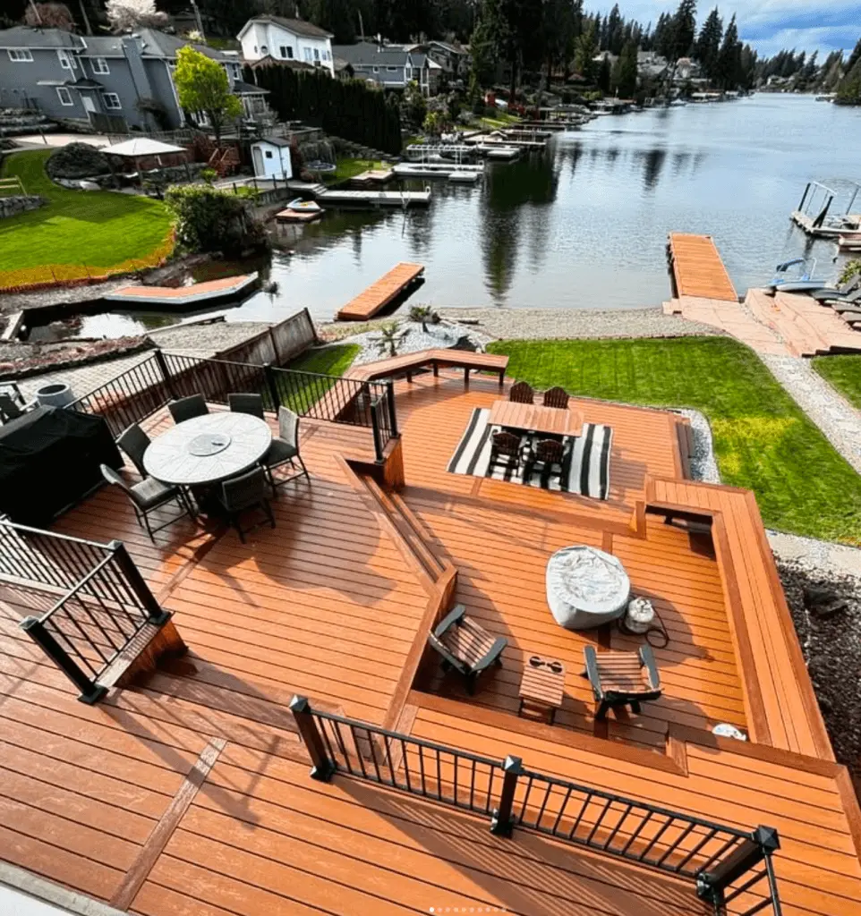 This is an example of our client's work. A multi-level wooden deck with seating areas, dining table, and fire pit overlooking a calm lake with private docks and surrounding houses.