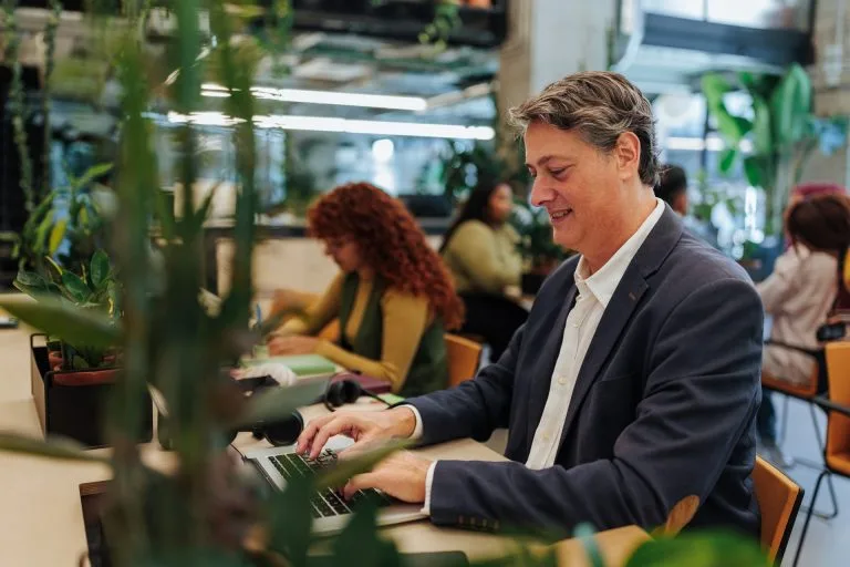 Smiling man in a blazer working on a laptop at a shared workspace, with plants and other people in the background.